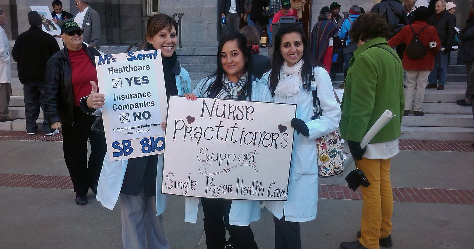 Three young women in lab coats holding up signs that say "Nurse Practitioners Support Single Payer Health Care"